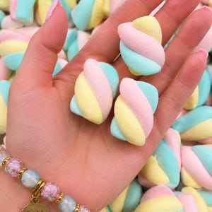 May include: Three twisted marshmallow candies featuring pastel pink, yellow, and blue stripes. The candies are held in a palm, with a beaded bracelet featuring a gold-toned circular charm engraved with the text Annie's Treasures and a small heart.