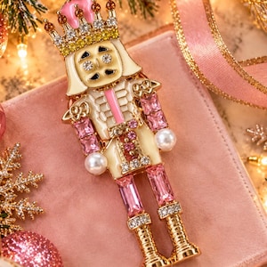 May include: A gold-tone nutcracker brooch featuring pink and cream enamel, accented with clear and pink rhinestones, and two white faux pearls. The figure wears a crown with pink and yellow stones. The text Annie's Treasures is at the bottom.