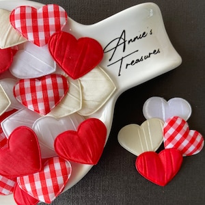 May include: A white ceramic dish with the text "Annie's Treasures" on it, filled with red and white gingham heart-shaped fabric patches. There are also solid red and white heart-shaped fabric patches in the dish.  There are three heart-shaped fabric patches on a black surface.
