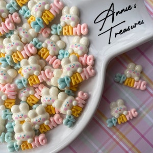 May include: A white plate filled with colorful resin bunny charms with the word "SPRING" written on them. The charms are in various colors, including pink, blue, yellow, and white. The plate is on a pink and white checkered background. The text "Annie's Treasures" is written on the plate.