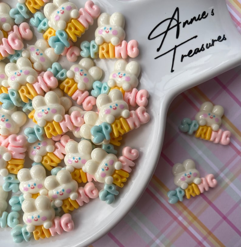 May include: A white plate filled with colorful resin bunny charms with the word "SPRING" written on them. The charms are in various colors, including pink, blue, yellow, and white. The plate is on a pink and white checkered background. The text "Annie's Treasures" is written on the plate.