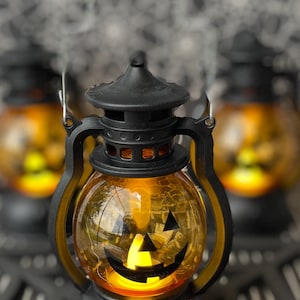 May include: A black and orange Halloween lantern with a smiling jack-o'-lantern face. The lantern has a handle and sits on a black base. An orange plastic spider is in the foreground. Other lanterns are in the background.