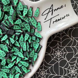 May include: A white ceramic dish filled with green and black poison bottle-shaped charms. Each charm features a skull and crossbones symbol. The dish is on a black and white spiderweb patterned surface. The text "Annie's Treasures" is written on the dish.
