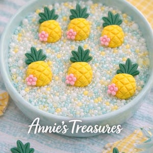 May include: A light blue bowl filled with small white, yellow, and blue beads, and decorated with yellow pineapple-shaped ornaments with green leaves and pink flowers. The text "Annie's Treasures" is visible.