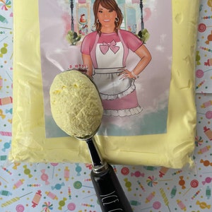 May include: A scoop of yellow ice cream in a silver ice cream scoop with a black handle that reads "ICE CREAM" in white. The ice cream is next to a bag of yellow frosting with a label that reads "Annie's Treasures". The background is a candy-themed pattern.