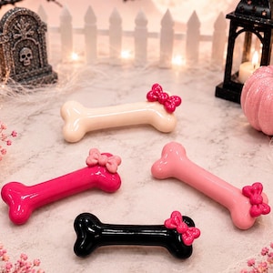 May include: Four smooth, glossy bone-shaped objects in cream, hot pink, light pink, and black. Each bone features a small, matching colored bow attached to one end. The text "Annie's Treasures" is printed in black script at the bottom of the image.