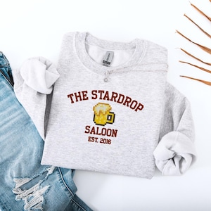 May include: A light gray sweatshirt with the text "THE STARDROP SALOON EST. 2016" in brown. The design includes a pixelated beer mug. The sweatshirt is paired with blue jeans.