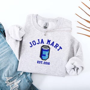 May include: A light grey sweatshirt featuring the blue text "JOJA MART" above a pixelated blue and white can graphic. Below the graphic is "EST. 2016" in blue. The sweatshirt is shown with light blue jeans.