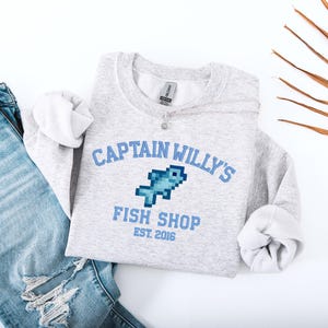 May include: A light grey sweatshirt featuring the text "CAPTAIN WILLY'S FISH SHOP EST. 2016" in blue, with a pixelated fish graphic. The sweatshirt is shown with light blue denim jeans.