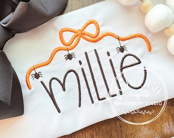 Pode incluir: Uma peça de tecido branco com um laço laranja bordado do qual pendem três pequenas aranhas pretas. Abaixo do laço, o nome Millie está bordado em letras cursivas castanho-escuras. Um grande laço de fita cinzento-escuro está fixado.