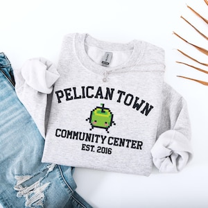 Könnte beinhalten: Hellgraues Sweatshirt mit dem schwarzen Schriftzug "PELICAN TOWN COMMUNITY CENTER EST. 2016". Darunter befindet sich eine pixelige grüne Apfelgrafik. Links ist eine hellblaue Jeans zu sehen.