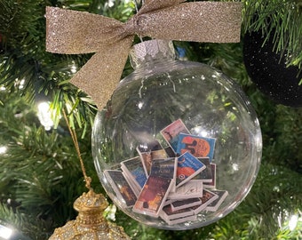 May include: A clear glass Christmas ornament with a gold glitter bow hanging on a green Christmas tree. Inside the ornament are miniature book covers.
