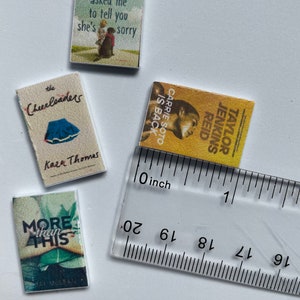 Puede incluir: Cuatro libros de bolsillo en miniatura con diferentes portadas. Los libros est&aacute;n dispuestos en fila sobre una superficie blanca. Una regla se coloca debajo de los libros. La regla est&aacute; marcada en pulgadas.