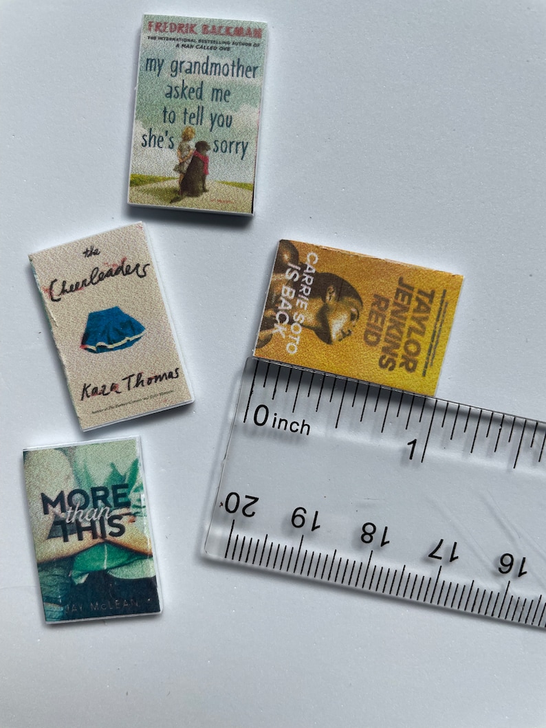 Puede incluir: Cuatro libros de bolsillo en miniatura con diferentes portadas. Los libros est&aacute;n dispuestos en fila sobre una superficie blanca. Una regla se coloca debajo de los libros. La regla est&aacute; marcada en pulgadas.