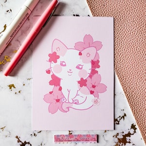 K&ouml;nnte beinhalten: Eine rosa und wei&szlig;e Illustration einer Katze, die in einem Bl&uuml;tenmeer aus rosa Kirschbl&uuml;ten sitzt. Die Katze hat eine rosa Nase und rosa Wangen. Die Illustration ist auf rosa Hintergrund.