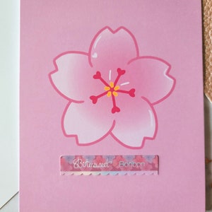 May include: A pink card with a hand-drawn pink cherry blossom flower in the center. The flower has a yellow center and red accents. The card has a small sticker with the text "Bittersweet Bonbon" at the bottom.
