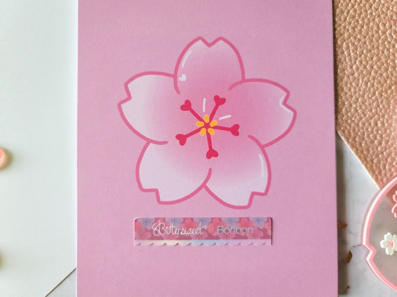 May include: A pink card with a hand-drawn pink cherry blossom flower in the center. The flower has a yellow center and red accents. The card has a small sticker with the text "Bittersweet Bonbon" at the bottom.