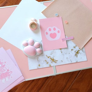 K&ouml;nnte beinhalten: Ein rosa Tisch mit einer Vielzahl von Bastelartikeln, darunter eine rosa Pfotenabdruck-Schablone, ein rosa Pfotenabdruck-Stempel, eine Rolle rosa Washi-Tape, ein rosa-wei&szlig;es Marmorpapier, ein rosa St&uuml;ck Kunstleder und eine rosa Karte mit einem rosa Pfotenabdruck-Design.