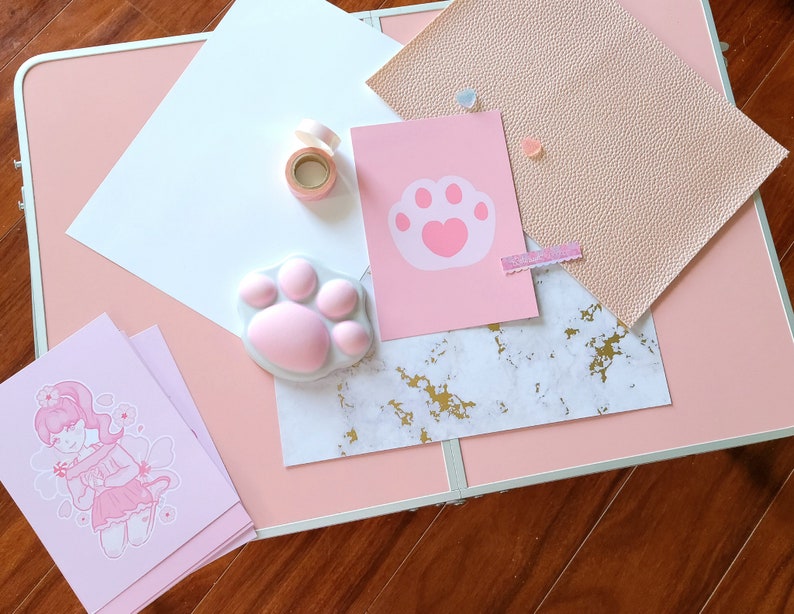 K&ouml;nnte beinhalten: Ein rosa Tisch mit einer Vielzahl von Bastelartikeln, darunter eine rosa Pfotenabdruck-Schablone, ein rosa Pfotenabdruck-Stempel, eine Rolle rosa Washi-Tape, ein rosa-wei&szlig;es Marmorpapier, ein rosa St&uuml;ck Kunstleder und eine rosa Karte mit einem rosa Pfotenabdruck-Design.