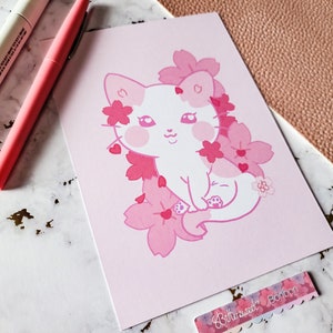K&ouml;nnte beinhalten: Eine rosa und wei&szlig;e Illustration einer Katze, die in einem Feld mit rosa Kirschbl&uuml;ten sitzt. Die Katze hat eine rosa Nase und rosa Wangen. Die Illustration ist auf rosa Hintergrund.