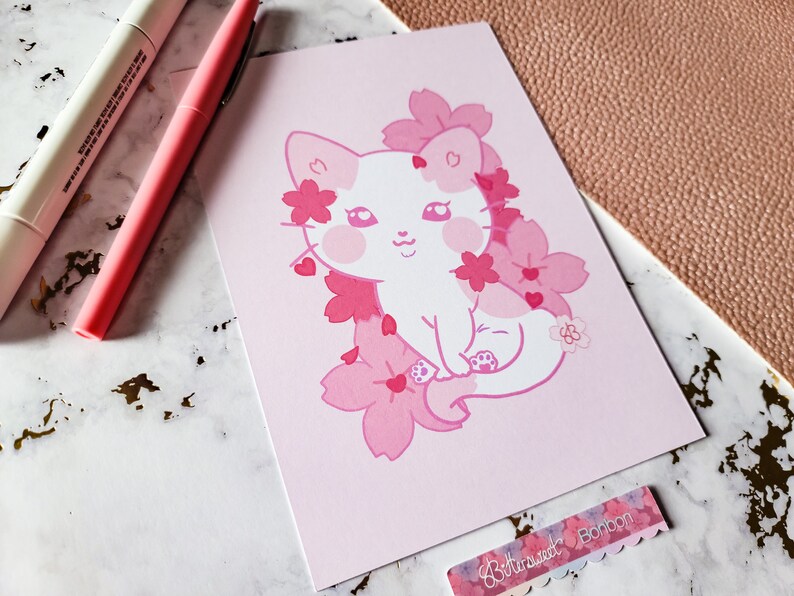 K&ouml;nnte beinhalten: Eine rosa und wei&szlig;e Illustration einer Katze, die in einem Feld mit rosa Kirschbl&uuml;ten sitzt. Die Katze hat eine rosa Nase und rosa Wangen. Die Illustration ist auf rosa Hintergrund.
