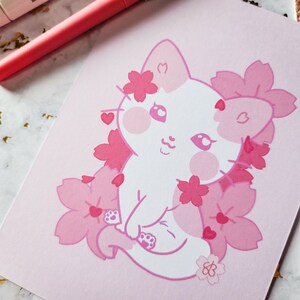 K&ouml;nnte beinhalten: Eine wei&szlig;e Katze mit rosa Wangen und einer rosa Nase ist von rosa Kirschbl&uuml;ten umgeben. Die Katze hat ein rosa Herz am Ohr und ein rosa Herz am Schwanz. Der Hintergrund ist rosa.