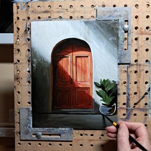 May include: A painting of a doorway with a red wooden door and a potted plant. The door is set in a white wall with a dark green archway. The painting is on an easel and is being worked on by an artist.