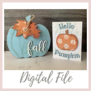 May include: A teal wooden pumpkin ornament with the word "fall" in white script, decorated with orange and brown paper leaves and twine. Beside it is a small white sign featuring a pumpkin and the words "Hello Pumpkin".