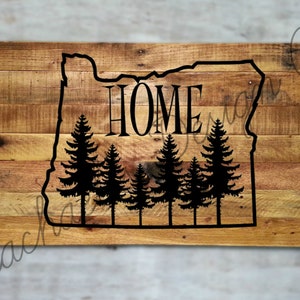 May include: Wooden sign featuring the outline of Oregon with the word "HOME" in the center. Below the word are silhouettes of pine trees. The sign is made of wooden planks.