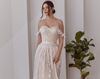 May include: A floor-length dress featuring a white lace overlay with intricate floral patterns over a beige lining. The garment has thin spaghetti straps, off-the-shoulder lace sleeves, a sweetheart neckline, and a thin white ribbon at the waist.