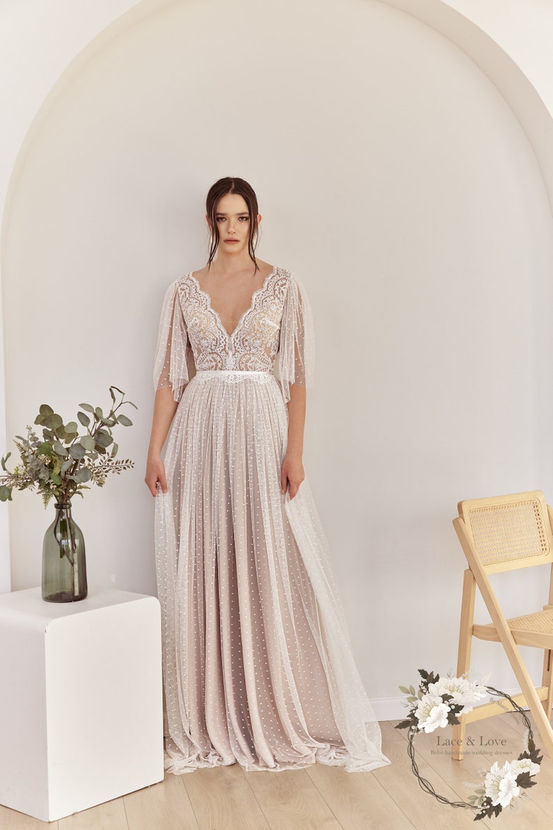 An ivory lace wedding dress with a v-neckline and a fitted bodice. The skirt is made of a sheer fabric with polka dots and has a flowing silhouette. The dress has a belt at the waist and short sleeves. Lace & Love Bohemian Wedding Dresses.