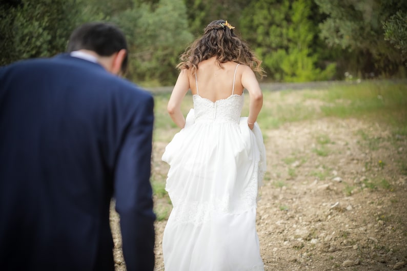 May include: A bride in a white wedding dress walks away from the groom, who is wearing a dark blue suit. The bride is wearing a flower crown in her hair.