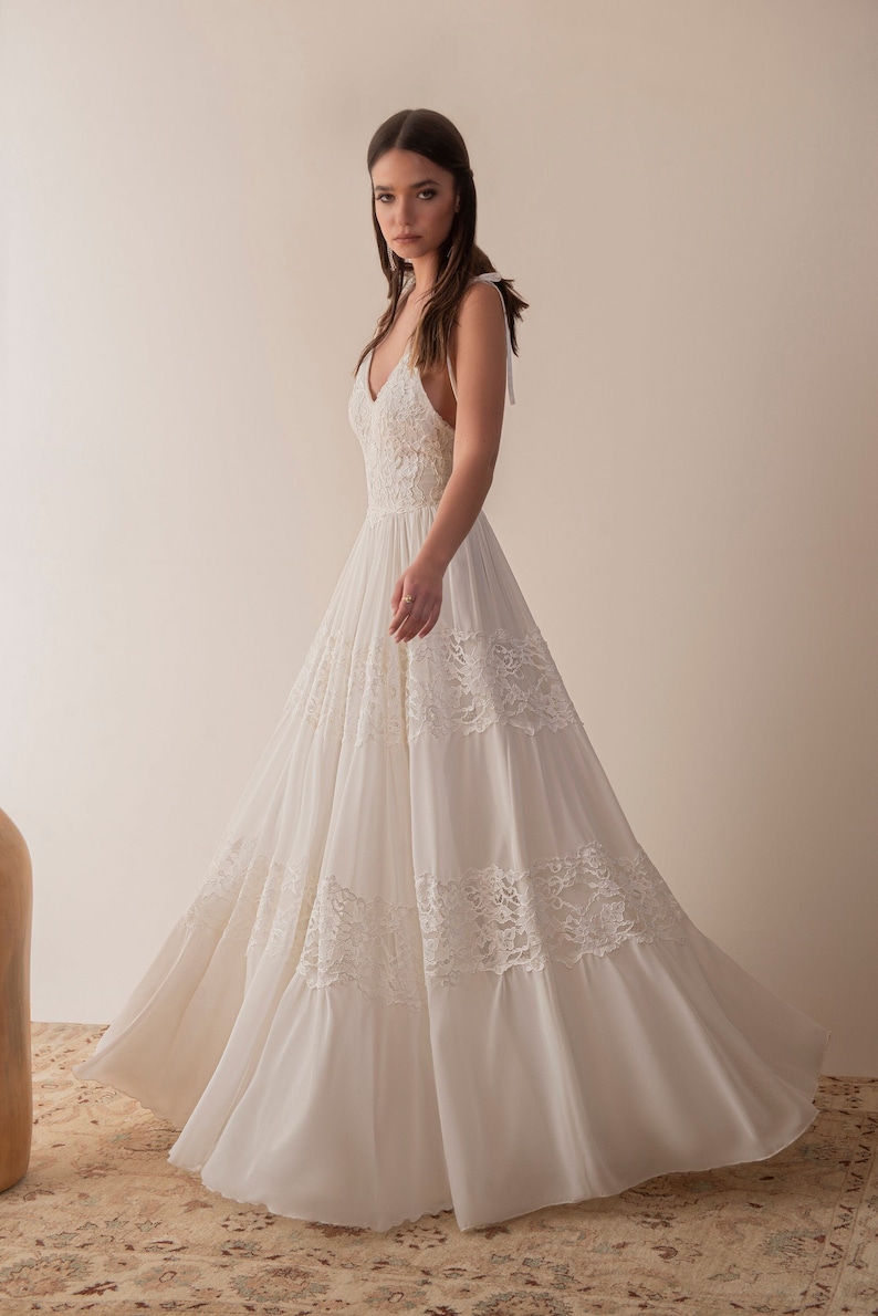 May include: A white sleeveless wedding dress with a lace bodice and a flowing skirt. The dress has a V-neckline and a tie at the back of the neck.