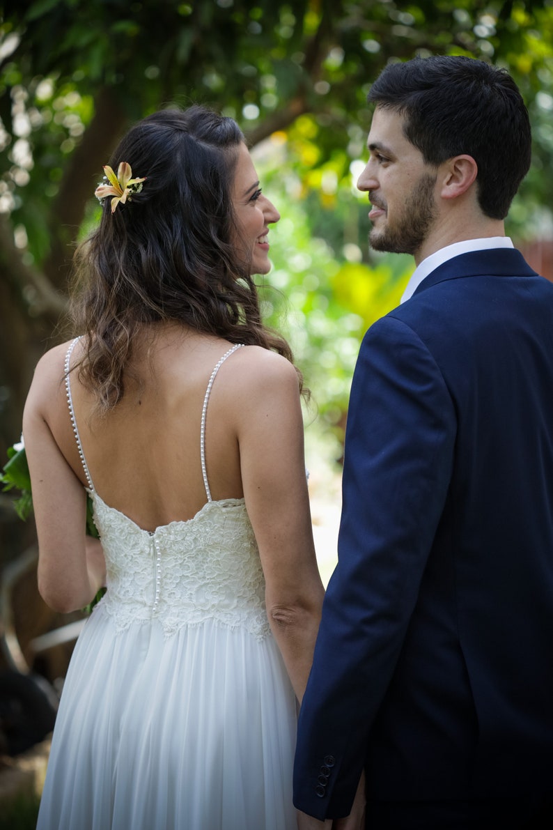 May include: A bride and groom stand facing each other in a garden setting. The bride is wearing a white lace wedding dress with a flower in her hair. The groom is wearing a dark blue suit.