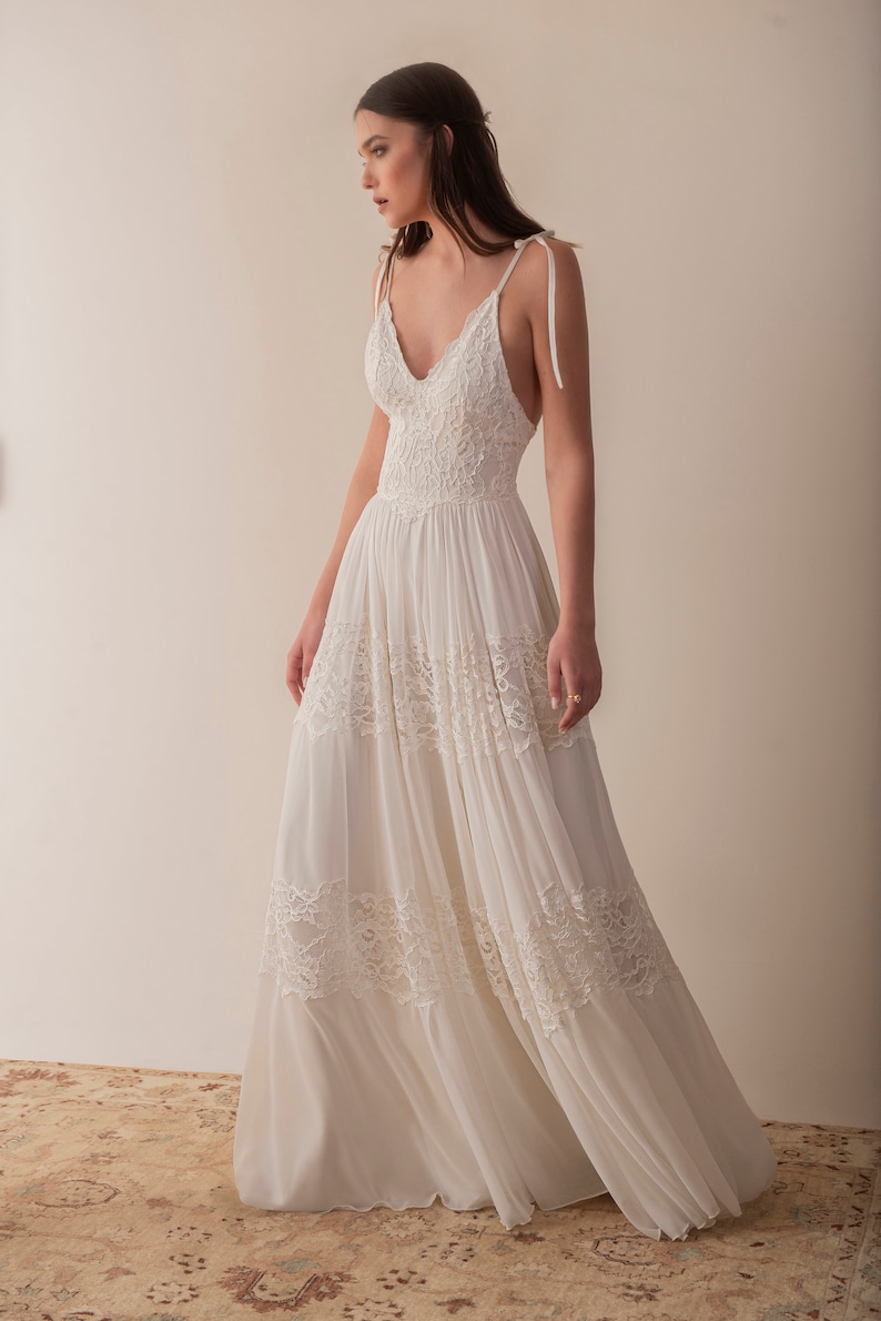 May include: A white sleeveless maxi dress with a lace bodice and a flowing skirt. The dress has a V-neckline and thin straps that tie at the shoulders.