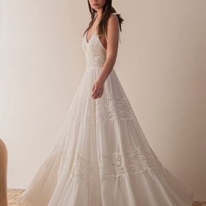 Puede incluir: Un vestido de novia blanco sin mangas con un corpi&ntilde;o de encaje y una falda fluida. El vestido tiene un escote en V y un lazo en la espalda.