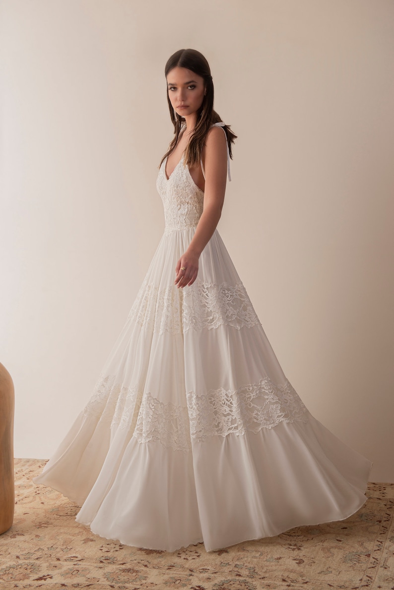 Puede incluir: Un vestido de novia blanco sin mangas con un corpi&ntilde;o de encaje y una falda fluida. El vestido tiene un escote en V y un lazo en la espalda.