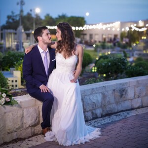 May include: A bride and groom are sitting on a stone wall, looking at each other. The bride is wearing a white wedding dress and the groom is wearing a blue suit. They are surrounded by greenery and a miniature city in the background.