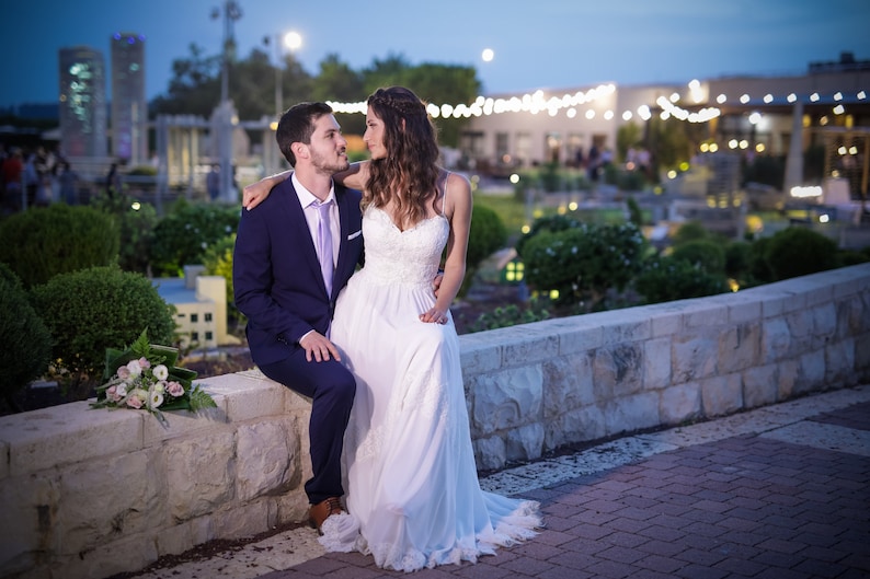 May include: A bride and groom are sitting on a stone wall, looking at each other. The bride is wearing a white wedding dress and the groom is wearing a blue suit. They are surrounded by greenery and a miniature city in the background.