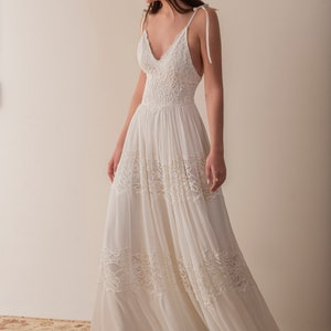 Puede incluir: Un vestido blanco maxi sin mangas con un corpi&ntilde;o de encaje y una falda fluida. El vestido tiene un escote en V y tirantes finos que se atan en los hombros.