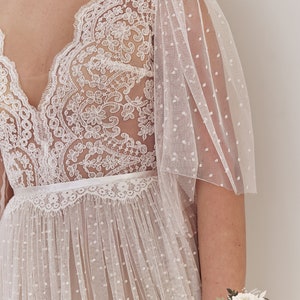 An ivory lace wedding dress with a v-neckline and a sheer, dotted tulle skirt. The dress has a delicate lace bodice and puffed sleeves. The sleeves are made of a sheer, dotted tulle fabric. The dress is accented with a white satin ribbon at the waist.