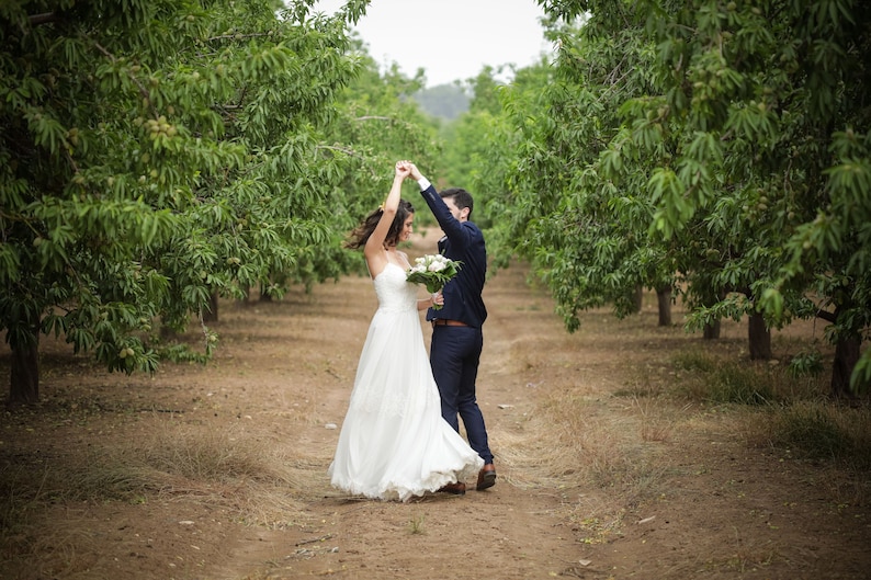 May include: A bride and groom in a wedding dress and suit dance in an orchard. The bride holds a bouquet of white flowers.