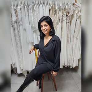 May include: A woman in a black shirt and black pants sits on a wooden stool in front of a wall of white lace wedding dresses. The sign above her reads "Lace & Love Brides - Made With Love - "