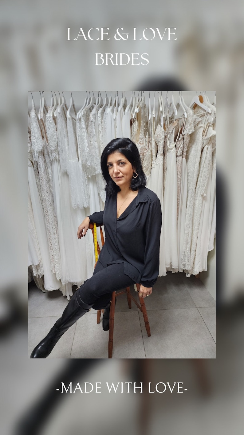 May include: A woman in a black shirt and black pants sits on a wooden stool in front of a wall of white lace wedding dresses. The sign above her reads "Lace & Love Brides - Made With Love - "