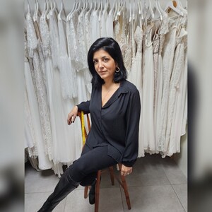 Puede incluir: Una mujer con una camisa negra y pantalones negros se sienta en un taburete de madera frente a una pared de vestidos de novia de encaje blanco. El letrero que est&aacute; encima de ella dice "Lace & Love Brides - Made with Love - "