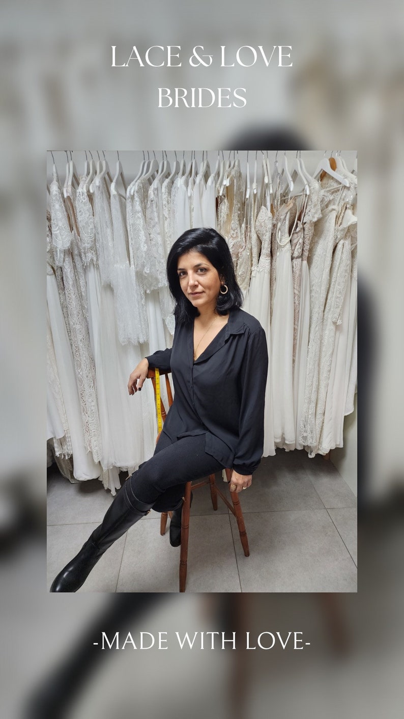Puede incluir: Una mujer con una camisa negra y pantalones negros se sienta en un taburete de madera frente a una pared de vestidos de novia de encaje blanco. El letrero que est&aacute; encima de ella dice "Lace & Love Brides - Made with Love - "