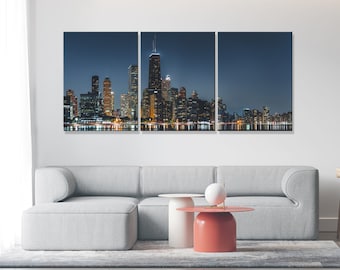 May include: A three-panel canvas print featuring a cityscape at night. The city skyline is illuminated with lights, reflecting in the water below. The print is displayed on a white wall above a gray couch and two coffee tables.