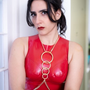 Puede incluir: Una mujer con un top rojo de látex con una cadena dorada y varios anillos. El top es sin mangas y tiene un escote redondo.