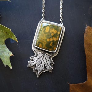 May include: A silver pendant necklace featuring a rectangular, speckled green and yellow stone set in a silver frame. The pendant is attached to a silver chain and has a leaf design at the bottom. The background includes green and brown leaves.