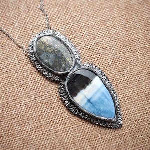 May include: A silver necklace with a pendant featuring two stone settings. The top stone is oval-shaped with gray and gold tones, while the bottom is a teardrop shape with blue, white, and black hues.
